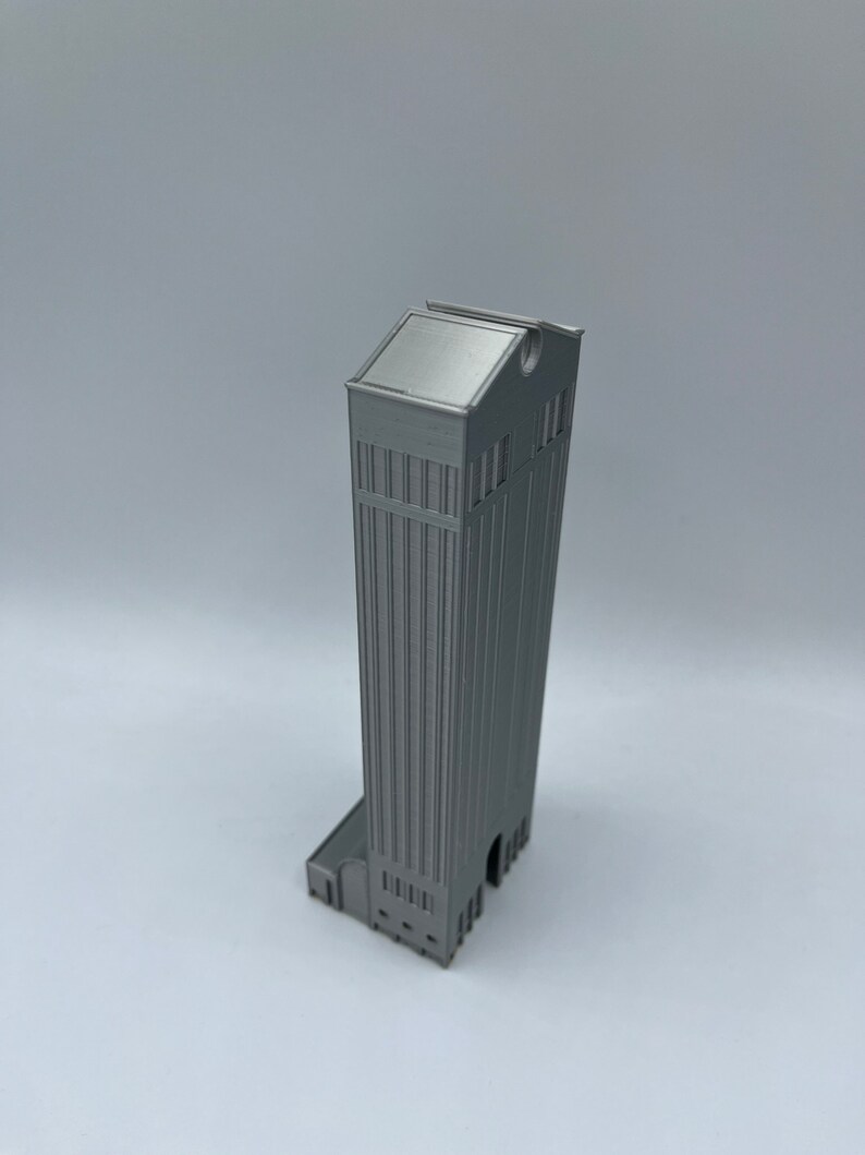 May include: A silver 3D printed model of a tall building with a flat roof and a rounded top. The building has a detailed facade with windows and a small entrance.