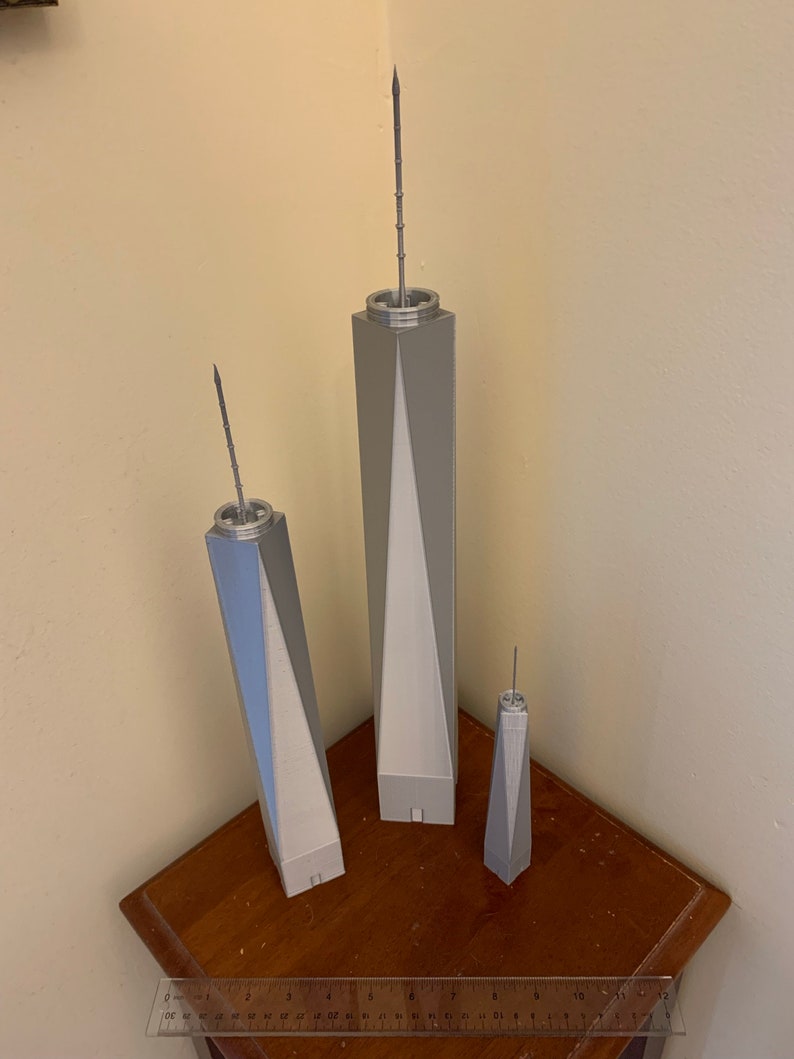 May include: Three silver model skyscrapers of varying sizes, the tallest is about 10 inches tall, the middle is about 6 inches tall, and the smallest is about 4 inches tall. The models are on a wooden surface with a ruler.