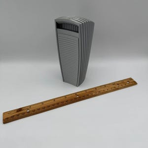May include: A gray, architectural-style sculpture with a curved, ribbed design. A wooden ruler, marked in inches, is placed next to the sculpture. The sculpture appears to be approximately 6 inches tall.