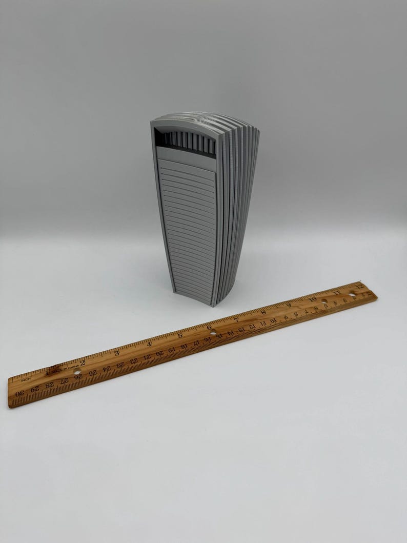 May include: A gray, architectural-style sculpture with a curved, ribbed design. A wooden ruler, marked in inches, is placed next to the sculpture. The sculpture appears to be approximately 6 inches tall.