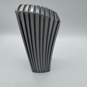 May include: A gray, abstract sculpture with a fan-like design. The object has a series of vertical, curved lines that create a ribbed texture. The sculpture is made of a solid material and stands on a white surface.