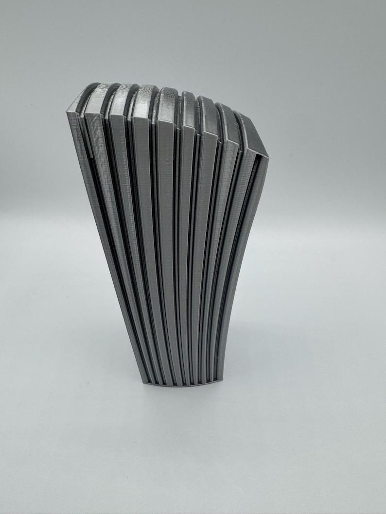 May include: A gray, abstract sculpture with a fan-like design. The object has a series of vertical, curved lines that create a ribbed texture. The sculpture is made of a solid material and stands on a white surface.