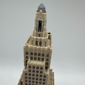 May include: A detailed, off-white miniature of a tall building with a gray, textured top. The building has many windows and architectural details, suggesting a skyscraper or historical structure. The model is likely 3D-printed.