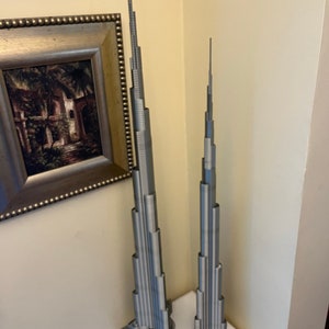 May include: Two silver metal models of the Burj Khalifa skyscraper, one larger than the other, on a white surface.