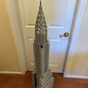 May include: A detailed model of the Chrysler Building in New York City, made of gray plastic. The model is tall and slender, with a pointed spire at the top. The building is decorated with intricate details, including windows, arches, and gargoyles.