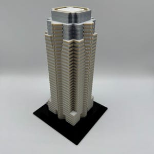 May include: A 3D-printed model of a skyscraper. The building is primarily white with gray and tan accents, and sits on a black square base. The top of the building has a flat roof.