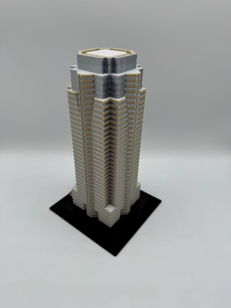 May include: A 3D-printed model of a skyscraper. The building is primarily white with gray and tan accents, and sits on a black square base. The top of the building has a flat roof.