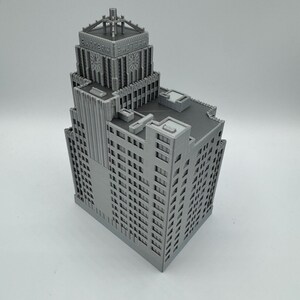 May include: A gray, 3D-printed model of a skyscraper. The building features detailed architectural elements, including numerous windows, vertical columns, and a decorative top with the text "EASTERN FACTORY". The model is a detailed representation of a classic building.