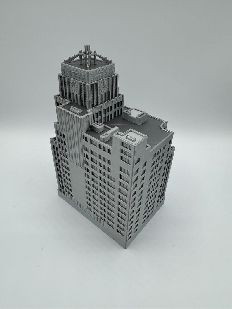 May include: A gray, 3D-printed model of a skyscraper. The building features detailed architectural elements, including numerous windows, vertical columns, and a decorative top with the text "EASTERN FACTORY". The model is a detailed representation of a classic building.