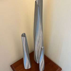 Op de afbeelding: Drie zilveren metalen vazen in verschillende maten, allemaal met een gedraaid, getextureerd ontwerp. De vazen staan op een houten tafel.