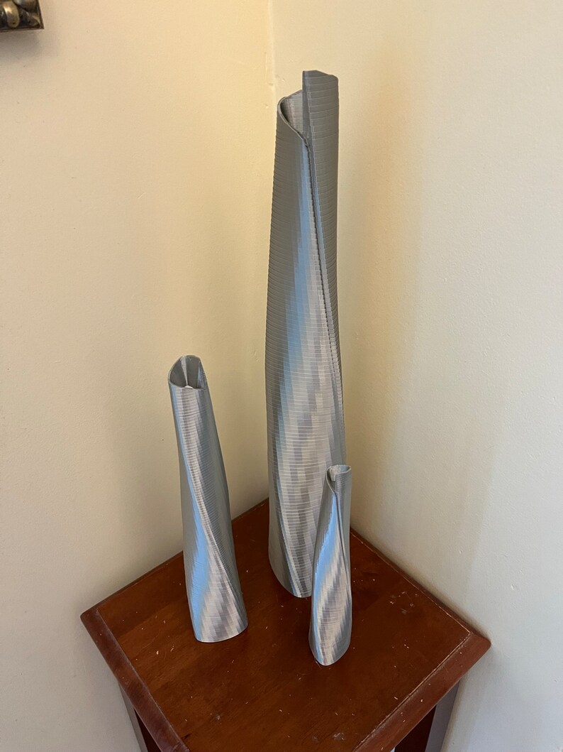 Op de afbeelding: Drie zilveren metalen vazen in verschillende maten, allemaal met een gedraaid, getextureerd ontwerp. De vazen staan op een houten tafel.