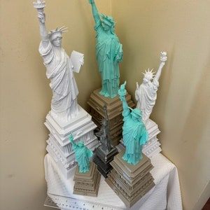 May include: A collection of five miniature Statue of Liberty figurines, each with a different base. The figurines are made of white and light blue plastic. The bases are made of brown plastic. The figurines are arranged on a white tablecloth.