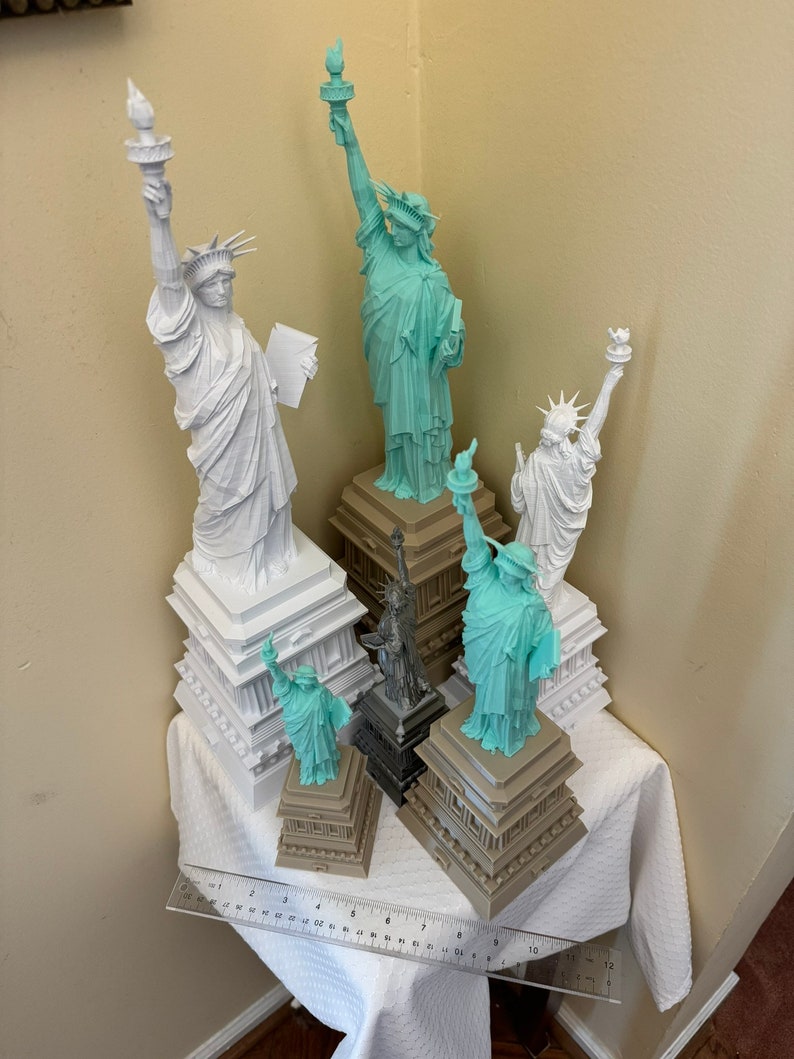 May include: A collection of five miniature Statue of Liberty figurines, each with a different base. The figurines are made of white and light blue plastic. The bases are made of brown plastic. The figurines are arranged on a white tablecloth.