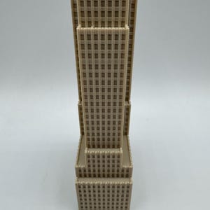 May include: A beige architectural model of a skyscraper. The building has a rectangular shape with multiple tiers and a grid-like pattern of windows. The model is made of a solid material and stands upright against a white background.