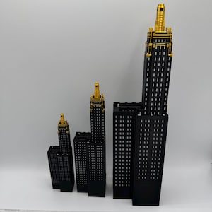 Op de afbeelding: Een verzameling zwarte miniatuur wolkenkrabbermodellen met goudkleurige toppen. De gebouwen vari&euml;ren in grootte en zijn in een groep gerangschikt. Een liniaal staat onderaan de afbeelding, met afmetingen in centimeters.