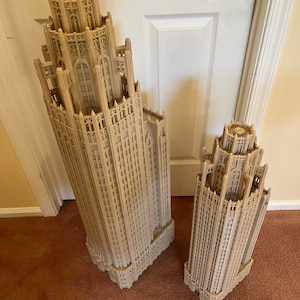 May include: Two detailed architectural models of skyscrapers, one larger than the other, are made of a light brown material and have a gothic design. The models are on a brown carpet.