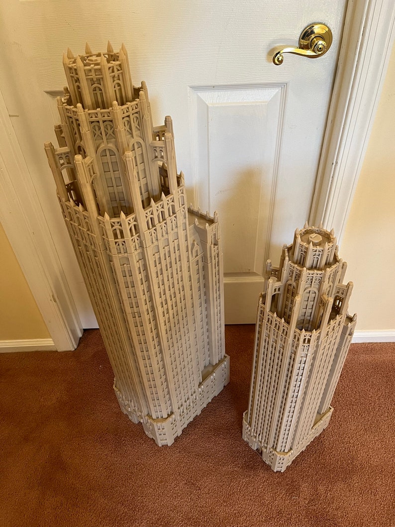 May include: Two detailed architectural models of skyscrapers, one larger than the other, are made of a light brown material and have a gothic design. The models are on a brown carpet.