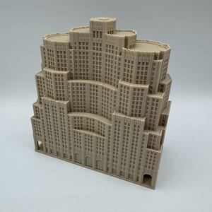 May include: A beige model of a multi-storey building with a stepped design. The model features many windows and architectural details, resembling a skyscraper. The building is made of a light-coloured material and sits on a white surface.