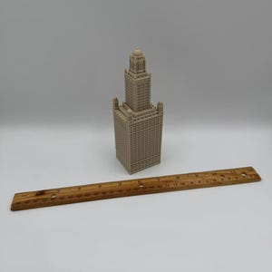 May include: A beige miniature replica of a tall building with a detailed facade. A wooden ruler, marked in inches, is placed next to the building. The building has a tower and multiple levels.