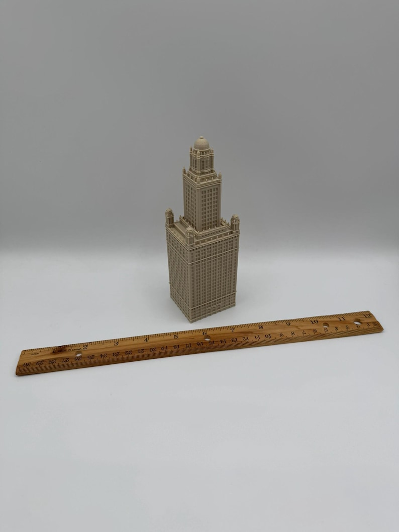 May include: A beige miniature replica of a tall building with a detailed facade. A wooden ruler, marked in inches, is placed next to the building. The building has a tower and multiple levels.