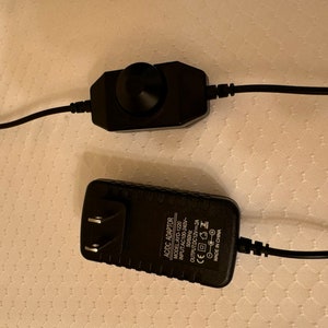 May include: A black AC adapter with a power cord and a black dimmer switch with a knob. The AC adapter has the text "AC/DC ADAPTOR MODEL AYD-1220 INPUT AC100-240V 50/60Hz OUTPUT DC12V 2A CE  MADE IN CHINA" printed on it.