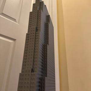 May include: A gray skyscraper model with a detailed facade featuring numerous windows. The building has a stepped design, with the top section narrower than the base. The model is positioned against a white door and a beige wall.