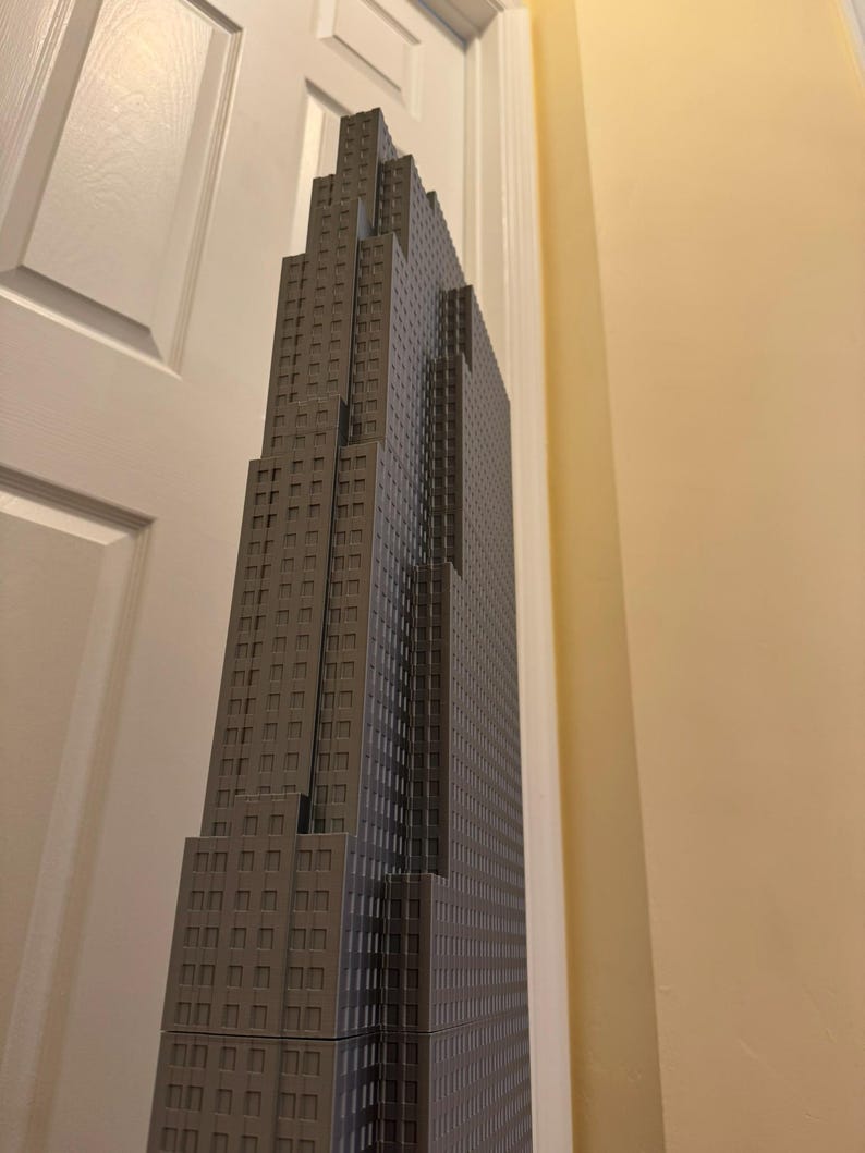May include: A gray skyscraper model with a detailed facade featuring numerous windows. The building has a stepped design, with the top section narrower than the base. The model is positioned against a white door and a beige wall.