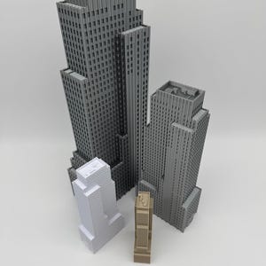 May include: A collection of miniature skyscraper models in varying sizes and colors, including gray, white, and tan. A wooden ruler is placed at the bottom of the image, providing a size reference. The buildings have detailed architectural features.