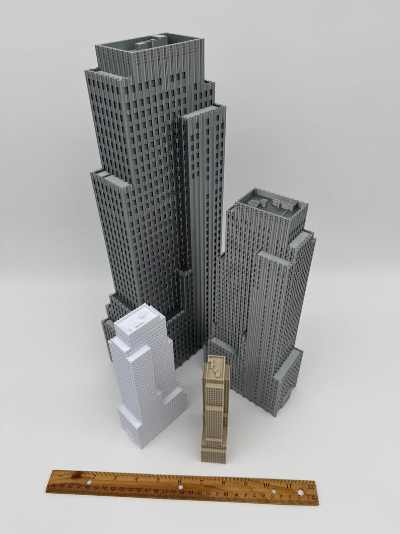 May include: A collection of miniature skyscraper models in varying sizes and colors, including gray, white, and tan. A wooden ruler is placed at the bottom of the image, providing a size reference. The buildings have detailed architectural features.