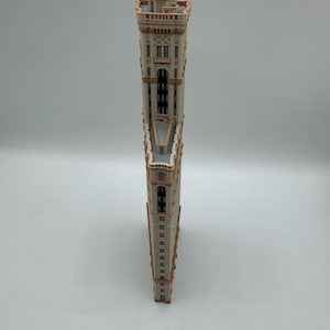May include: A wooden model of a tall building with a unique, triangular shape. The building has a beige facade with brown trim, windows, and architectural details. The model is standing upright against a white background.
