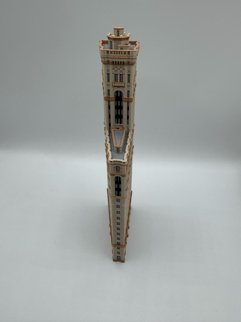 May include: A wooden model of a tall building with a unique, triangular shape. The building has a beige facade with brown trim, windows, and architectural details. The model is standing upright against a white background.