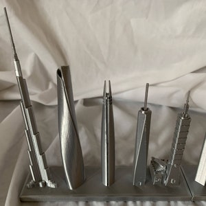 May include: A collection of silver-colored miniature skyscraper models. The models vary in design, with some featuring spires and others having unique architectural shapes. They are displayed on a gray rectangular base.