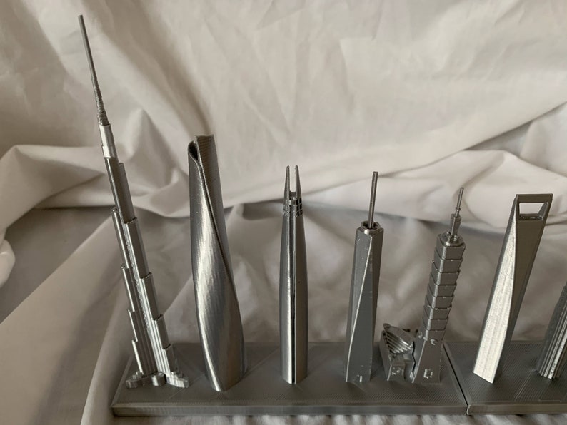May include: A collection of silver-colored miniature skyscraper models. The models vary in design, with some featuring spires and others having unique architectural shapes. They are displayed on a gray rectangular base.