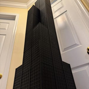 May include: A black architectural model of a skyscraper, with a rectangular design and multiple levels, featuring a textured facade. The model is set against a yellow wall and white doors.