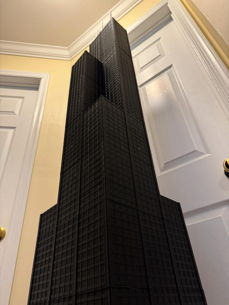May include: A black architectural model of a skyscraper, with a rectangular design and multiple levels, featuring a textured facade. The model is set against a yellow wall and white doors.
