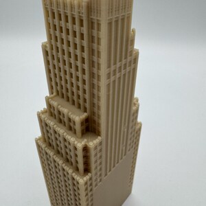 May include: A tan-colored, multi-tiered architectural model of a skyscraper. The model features detailed window and facade designs, with a rectangular base and a series of setbacks. The building's vertical lines and grid-like windows are prominent.