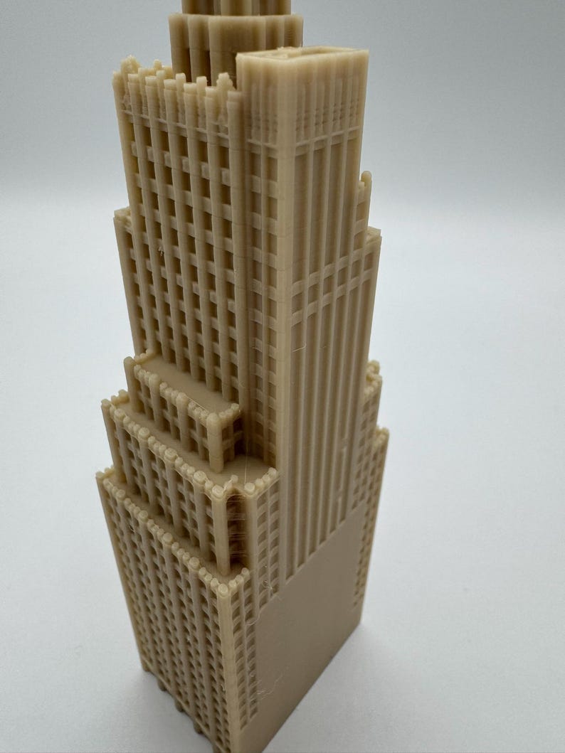May include: A tan-colored, multi-tiered architectural model of a skyscraper. The model features detailed window and facade designs, with a rectangular base and a series of setbacks. The building's vertical lines and grid-like windows are prominent.