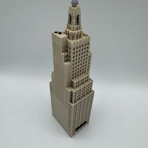 May include: A beige miniature skyscraper model with a detailed design. The building has multiple stories with rows of windows and a decorative top. The model is made of a light-colored material and stands upright against a white background.