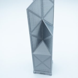May include: A gray, 3D-printed architectural model of a skyscraper. The building features a geometric design with triangular and rectangular shapes. The model is standing upright against a white background.