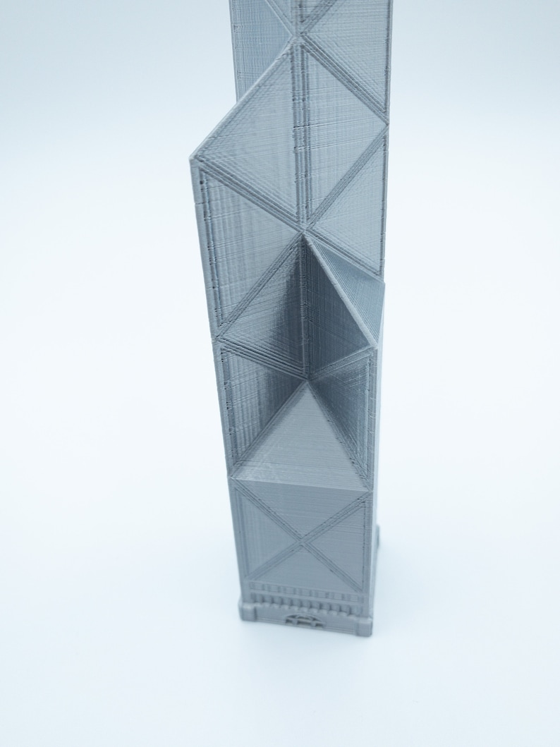 May include: A gray, 3D-printed architectural model of a skyscraper. The building features a geometric design with triangular and rectangular shapes. The model is standing upright against a white background.