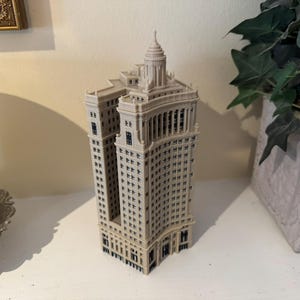 Op de afbeelding: Een miniatuurmodel van een hoog, beige gebouw met een afgeronde hoek en een decoratieve top. Het gebouw heeft veel ramen en is versierd met zwarte accenten.
