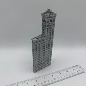 May include: A grey miniature model of a tall building with many windows and architectural details. A ruler with centimetre measurements is placed in front of the building, showing its scale. The building has a unique, angled design.