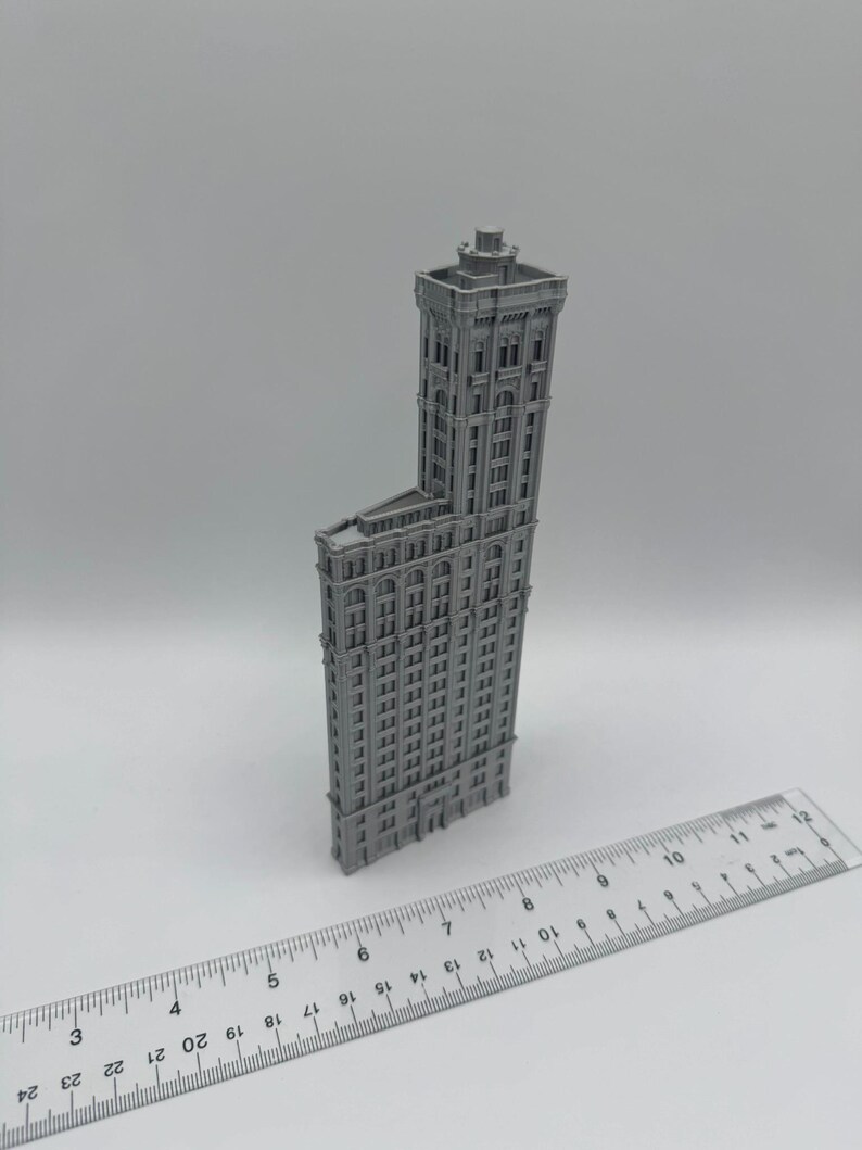 May include: A grey miniature model of a tall building with many windows and architectural details. A ruler with centimetre measurements is placed in front of the building, showing its scale. The building has a unique, angled design.