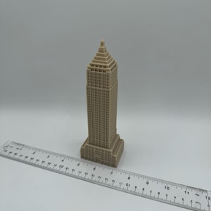 Puede incluir: Un modelo en miniatura beige de un rascacielos con un techo puntiagudo y muchas ventanas. El modelo mide aproximadamente 18 cm de alto y est&aacute; sentado sobre una superficie blanca junto a una regla.