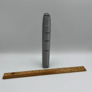 May include: A gray miniature skyscraper model stands upright next to a wooden ruler. The ruler is marked with inch measurements. The skyscraper model is a detailed replica of a tall building.