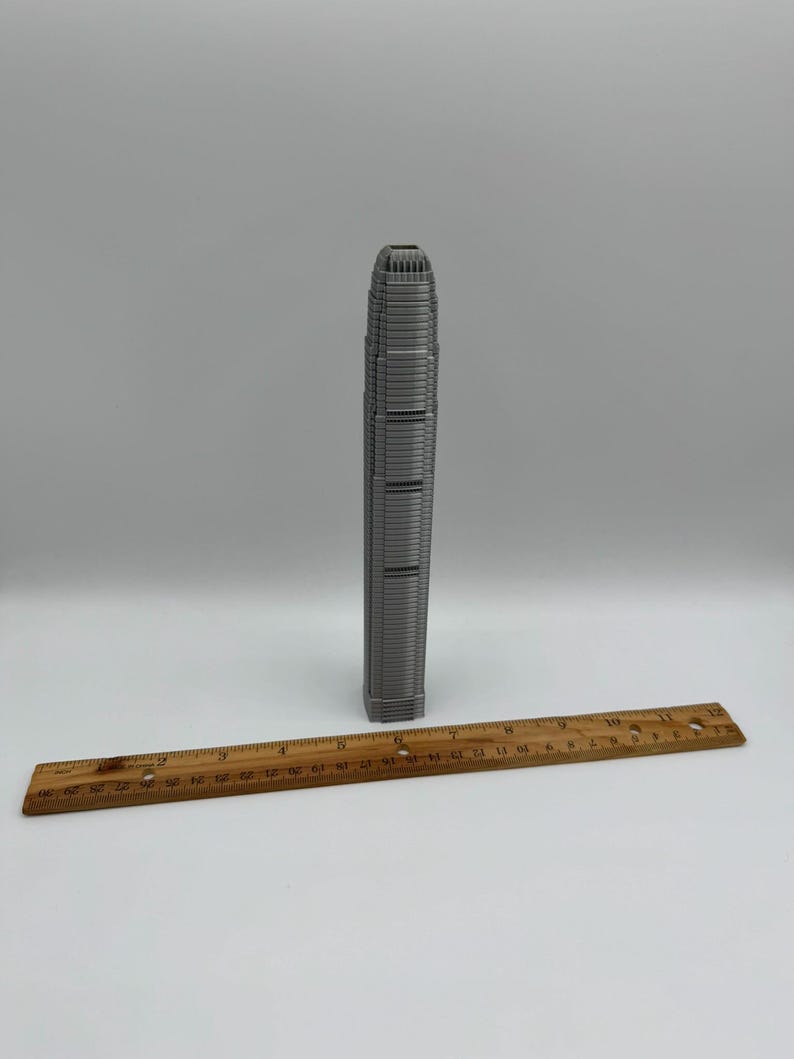 May include: A gray miniature skyscraper model stands upright next to a wooden ruler. The ruler is marked with inch measurements. The skyscraper model is a detailed replica of a tall building.