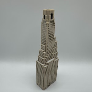 May include: A beige 3D printed architectural model of a skyscraper featuring a stepped, tiered design with a grid of small square windows covering the exterior facade. The structure has a tall, narrow central tower and a rectangular base.
