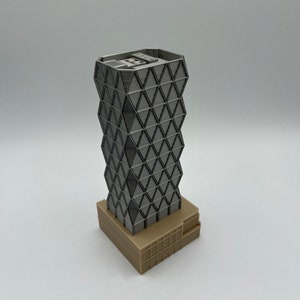 May include: A 3D printed model of a modern skyscraper with a geometric facade. The building is made of grey plastic and has a brown base.