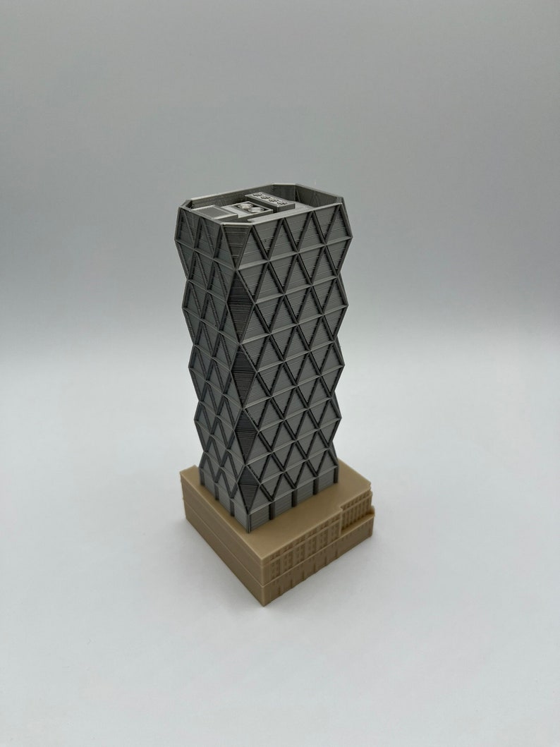May include: A 3D printed model of a modern skyscraper with a geometric facade. The building is made of grey plastic and has a brown base.