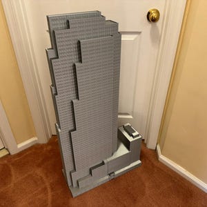 May include: A large, gray model of a skyscraper, featuring a stepped design and multiple levels. The model is made of small, textured blocks, and stands upright against a white door and a yellow wall. The model is approximately 4 feet tall.